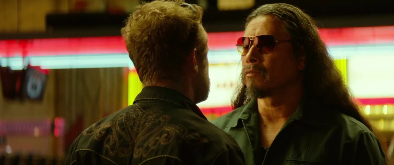 Hell or High Water (2016) movie still: medium — An outdoor space at night illuminated by the glow of bright red neon signs. Two men stand in close…
