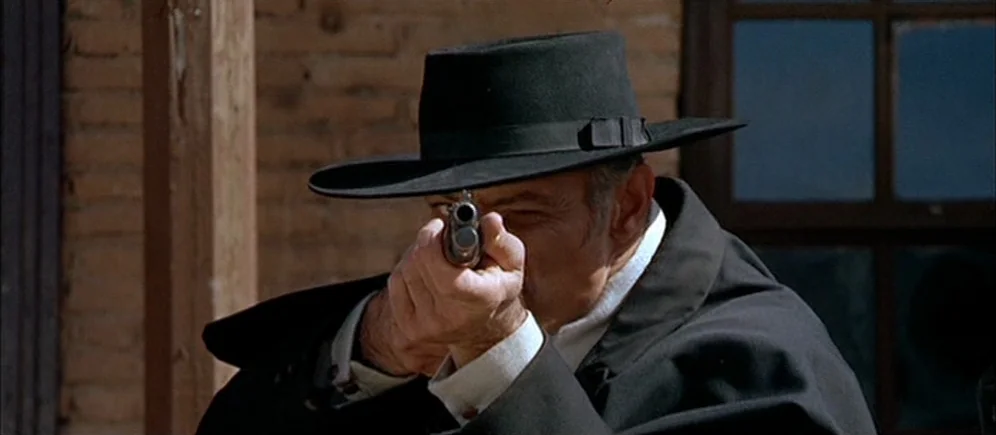 For A Few Dollars More (1965) movie still: closeup — A man in a black hat aims a handgun from behind at an unknown target. He is situated outdoors…