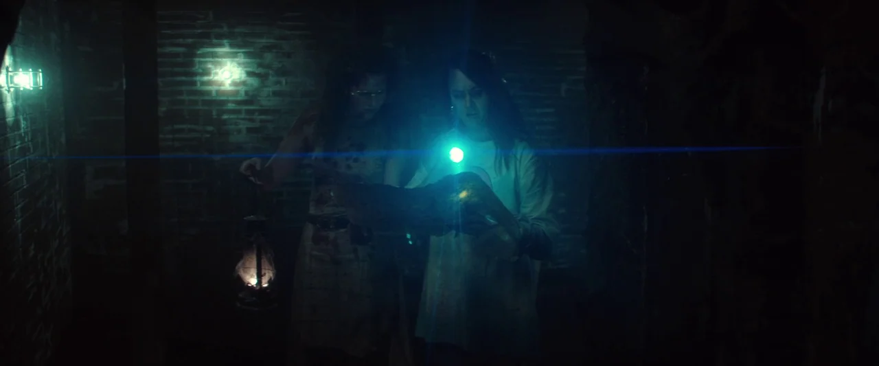 Revealer (2022) movie still: medium — A dark brick walled space features low-hanging pipes and dim ambient light sources. Two women stand together…