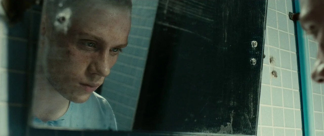 A Prayer Before Dawn (2017) movie still: closeup — A stark restroom with blue tiled walls and a damaged mirror featuring bullet holes. A man wearing…
