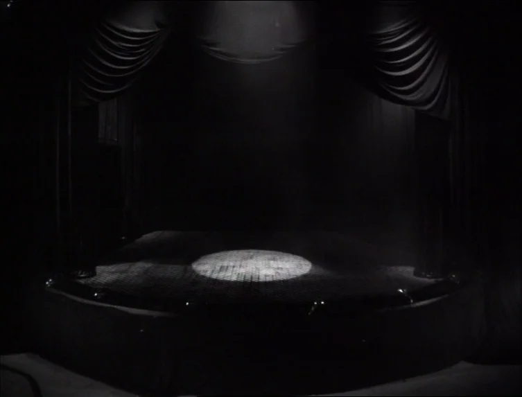 Eraserhead (1977) movie still: establishing — A dark empty performance area is framed by heavy black curtains on both sides. A circular spotlight…
