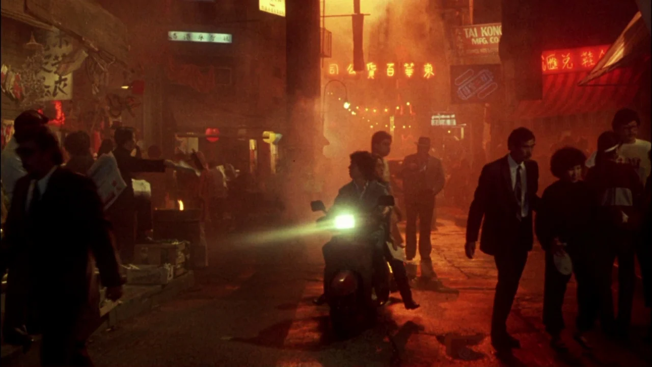 Gremlins (1984) movie still: wide — A crowded, smoke-filled street at night is illuminated by bright red and orange neon signage. People walk past a…