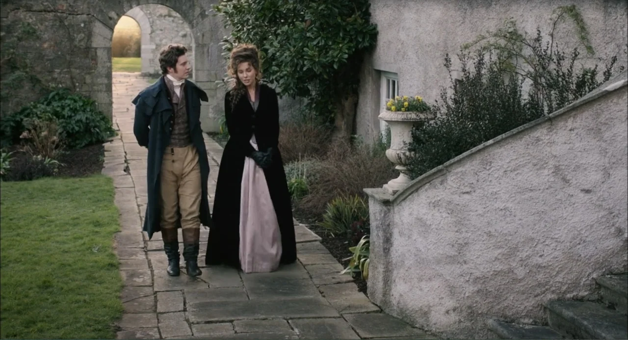 Love & Friendship (2016) movie still: fullbody — A stone walkway leads through a courtyard past a stone wall and stairs. A man and a woman in…
