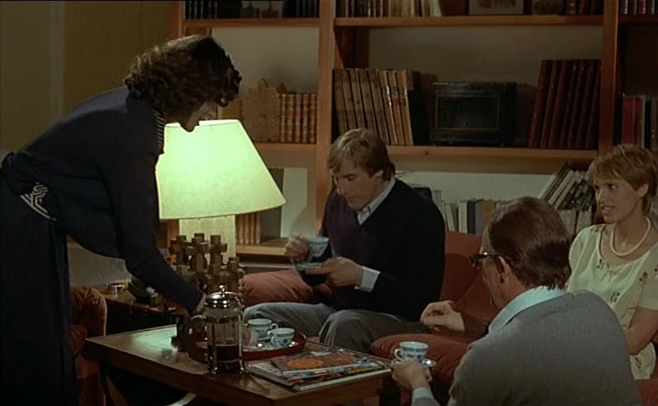 The Woman Next Door (1981) movie still: medium — A living room interior features a wooden bookshelf filled with books behind a seating area. A woman…
