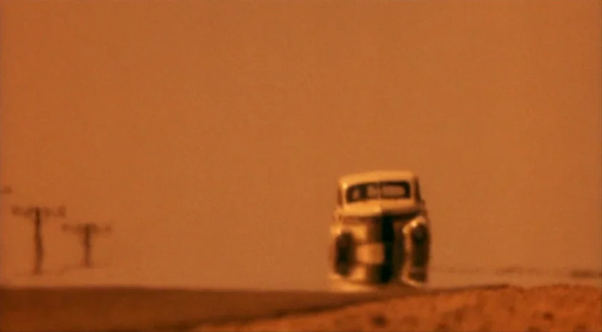 Dust Devil (1992) movie still: wide — A flat, orange-toned landscape stretches out under a haze with a lone vintage car. Two distant utility poles…