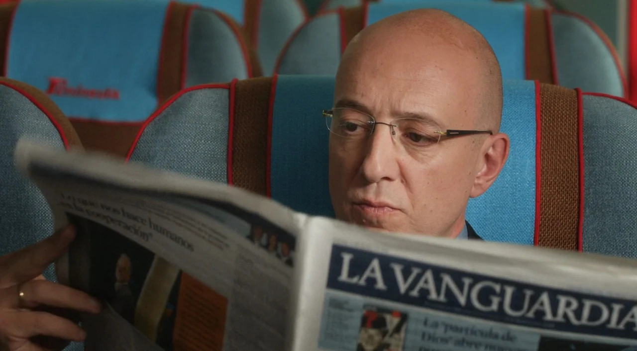 I’m So Excited! (2013) movie still: closeup — The interior of a passenger train features blue seats with red piping accents. A middle-aged bald man…