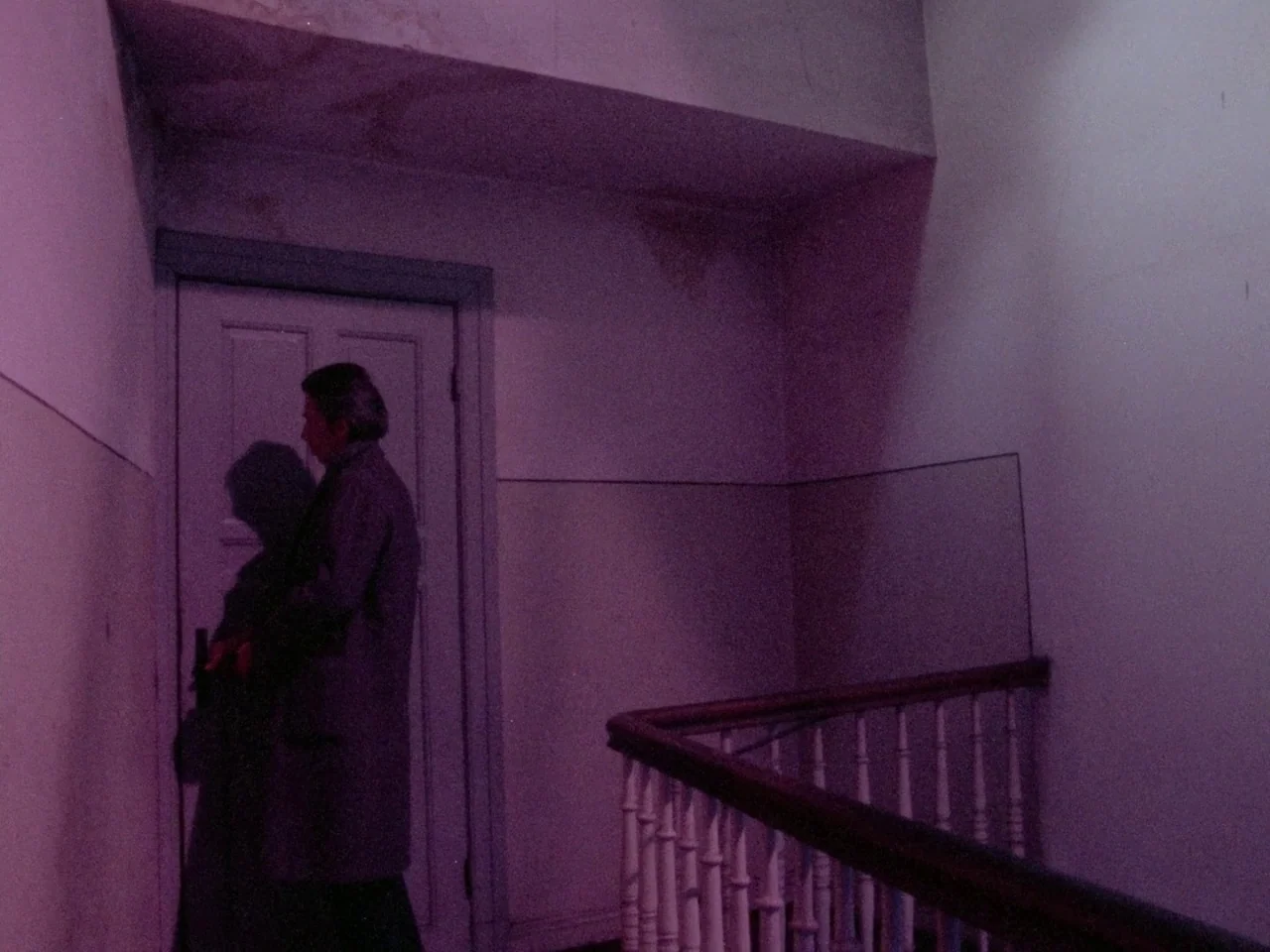 Decoder (1984) movie still: medium — A dimly lit stairwell landing features a closed door and a simple wooden banister. A man stands silently by the…