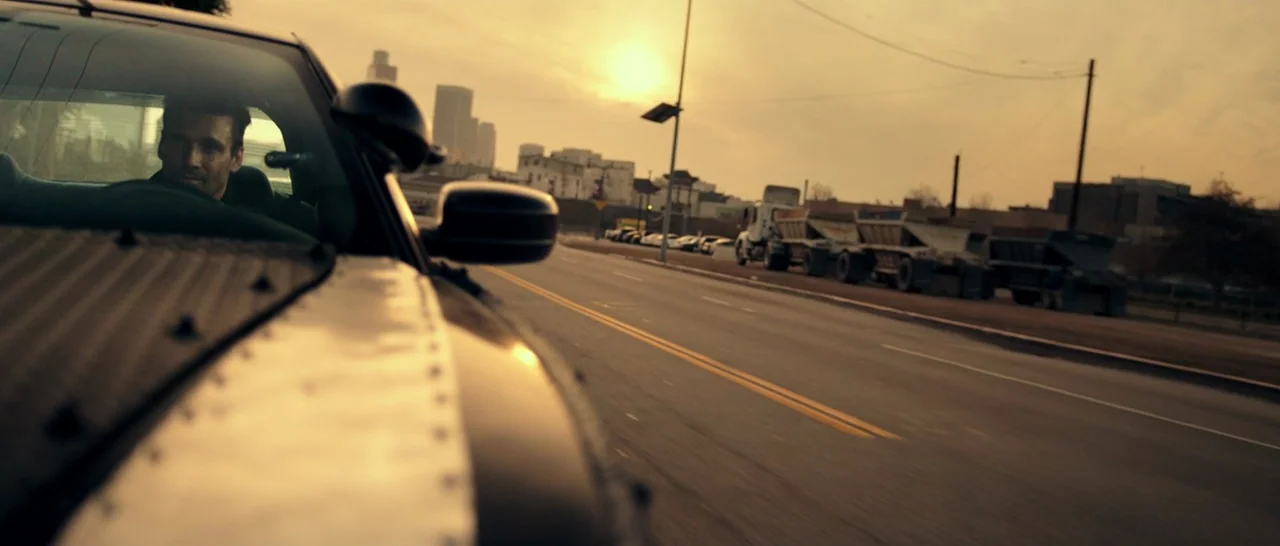 The Purge: Anarchy (2014) movie still: medium — A man drives a car along a road at golden hour with city buildings visible. The side of the vehicle…