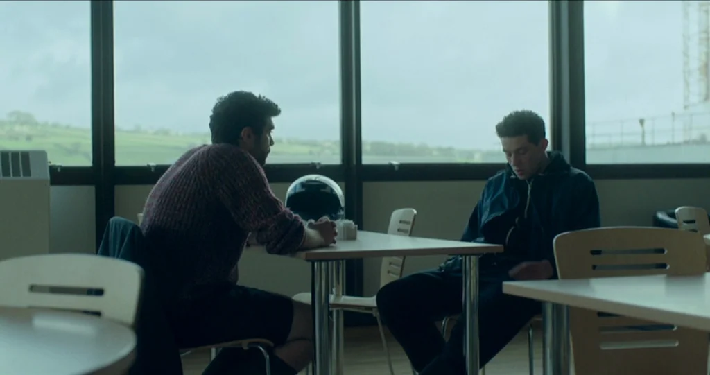 God’s Own Country (2017) movie still: medium — An interior cafeteria space features large windows overlooking a green landscape. Two men sit at a…