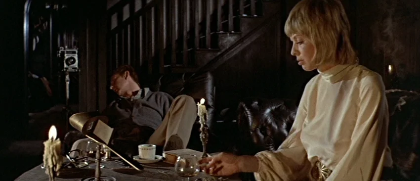 Images (1972) movie still: medium — A dimly lit room features a wooden table with a melting candle and drinks. A man rests in a leather armchair as…