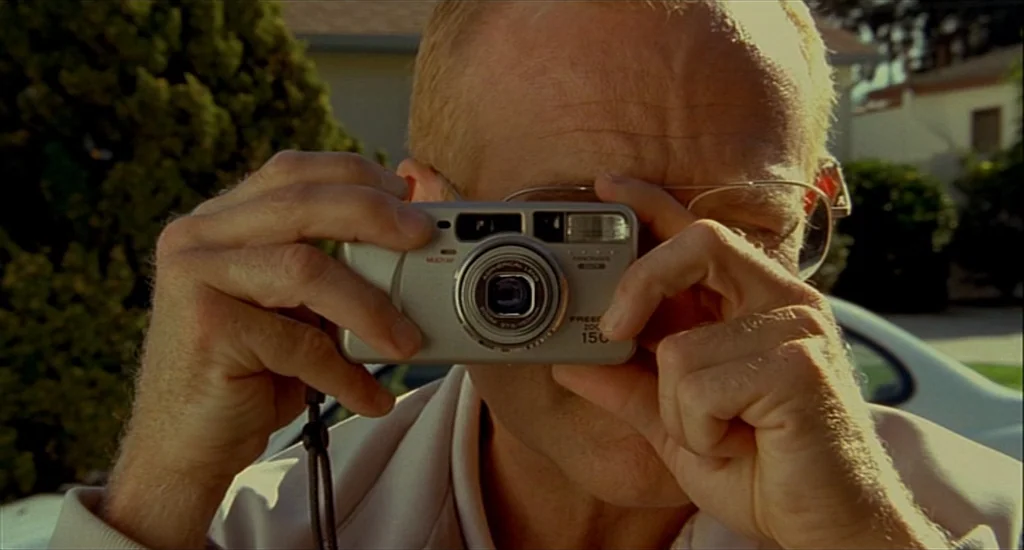 One Hour Photo (2002) movie still: closeup — A man in a suburban residential area stands outdoors near a white car. The man holds a silver compact…