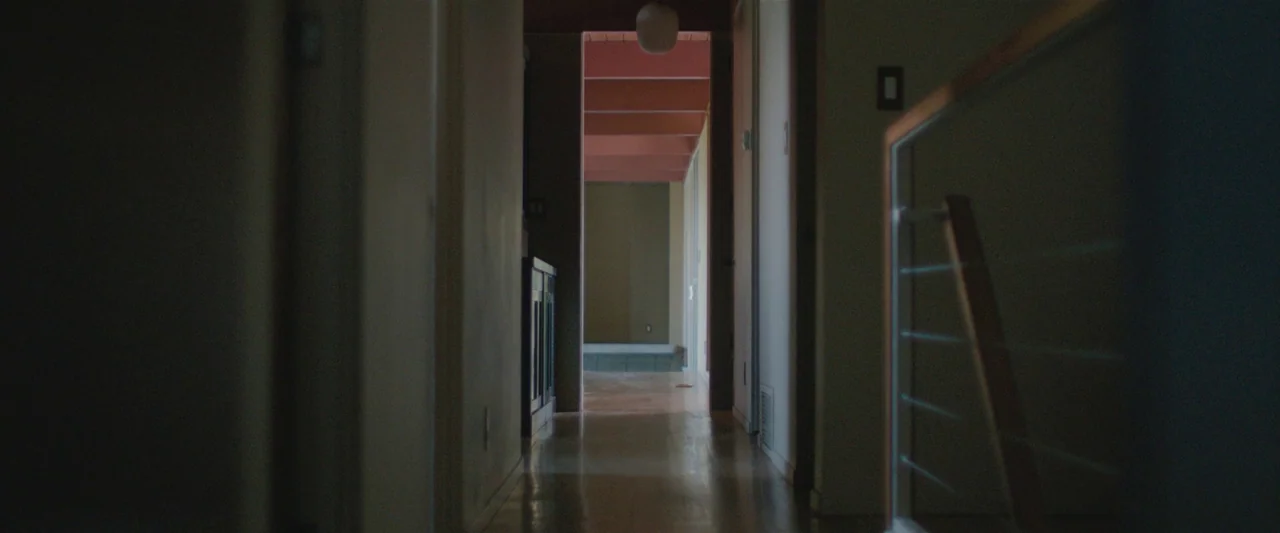 The Gift (2015) movie still: establishing — A dimly lit narrow residential hallway leads toward a brighter room with pink ceiling beams. The hallway…