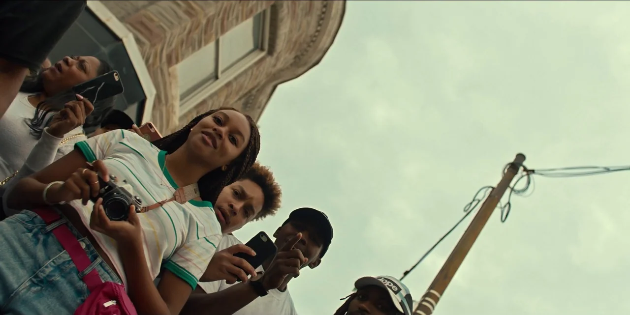 Charm City Kings (2020) movie still: medium — A group of young people stands on a city street under a cloudy sky. The subjects look down at the…
