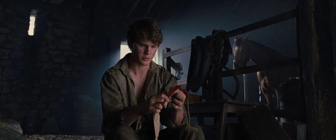 War Horse (2011) movie still: medium — A dimly lit barn interior features stone walls and wooden structures. A young man sits holding a piece of red…