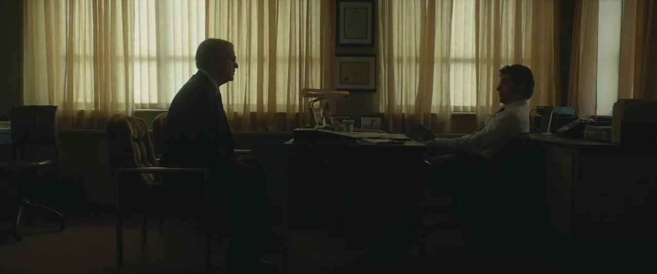 A Most Violent Year (2014) movie still: wide — A dimly lit office features two men sitting across a desk near large windows. The silhouettes of the…