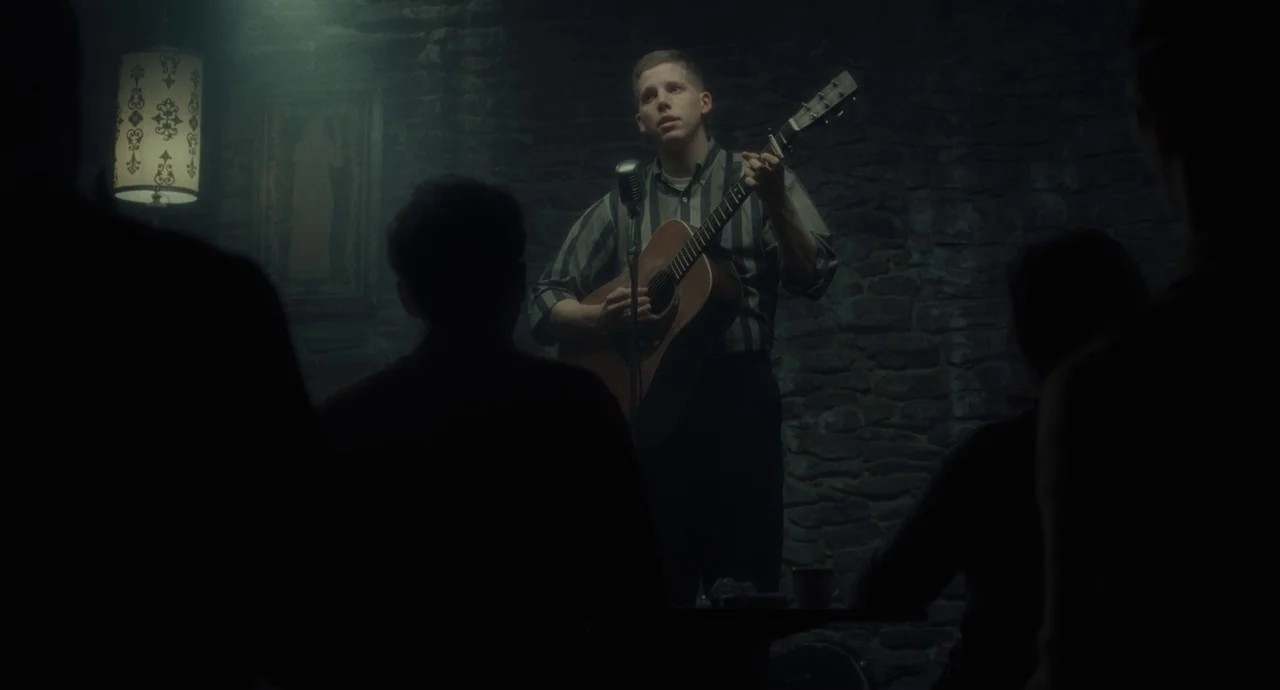 Inside Llewyn Davis (2013) movie still: medium — A dimly lit room with dark stone walls and a warm hanging lamp. A young man performs on stage with…