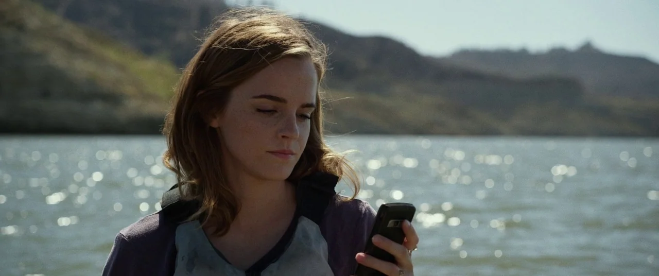 The Circle (2017) movie still: medium — A woman stands in front of a wide, sunlit lake with distant brown mountains. She holds a black smartphone…
