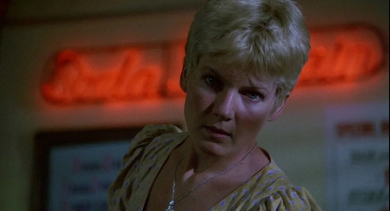 Psycho III (1986) movie still: closeup — A woman with short blonde hair stands inside a dimly lit diner interior. She looks toward the camera with…