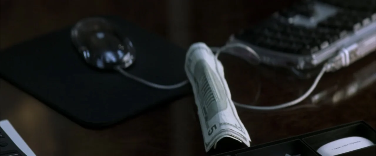 Enough (2002) movie still: closeup — A dark wooden desk surface holds a computer mouse, a rolled five-dollar bill, and a keyboard. These objects are…