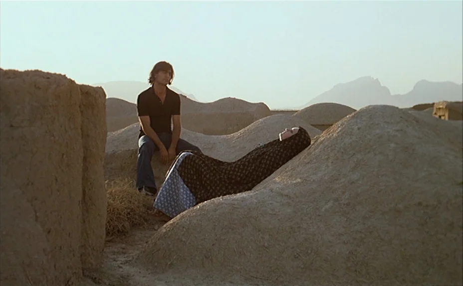 L’une Chante, l’Autre Pas (1977) movie still: wide — An arid, sandy landscape features undulating earthen mounds under a hazy sky. A man sits atop…