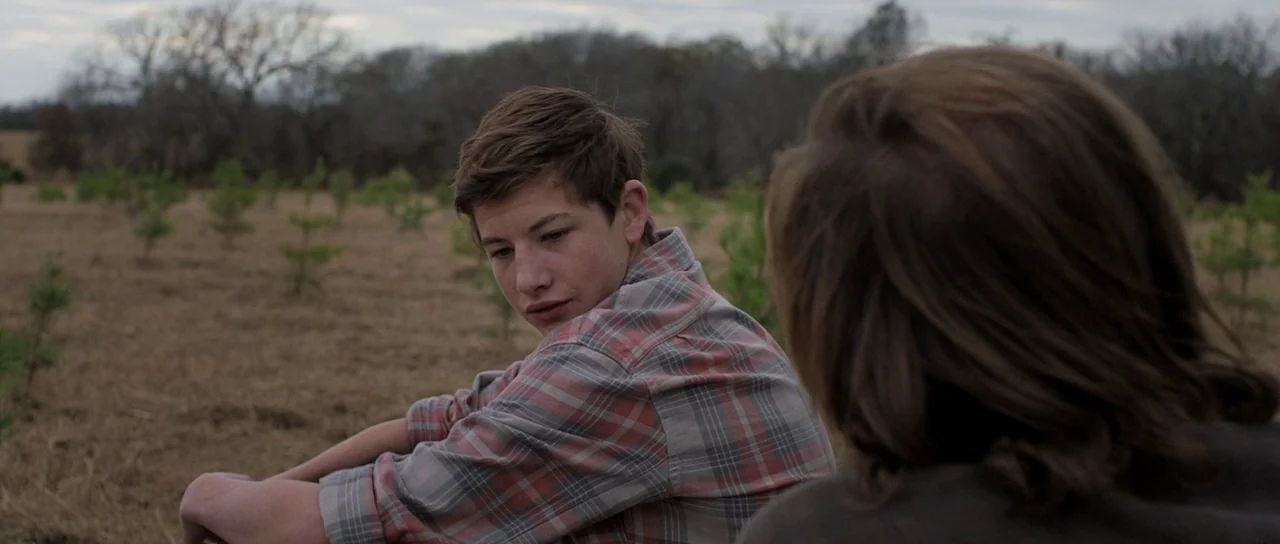 Joe (2013) movie still: medium — A field of dry grass and small saplings stretches toward a wooded horizon line. A young man sits in the foreground…