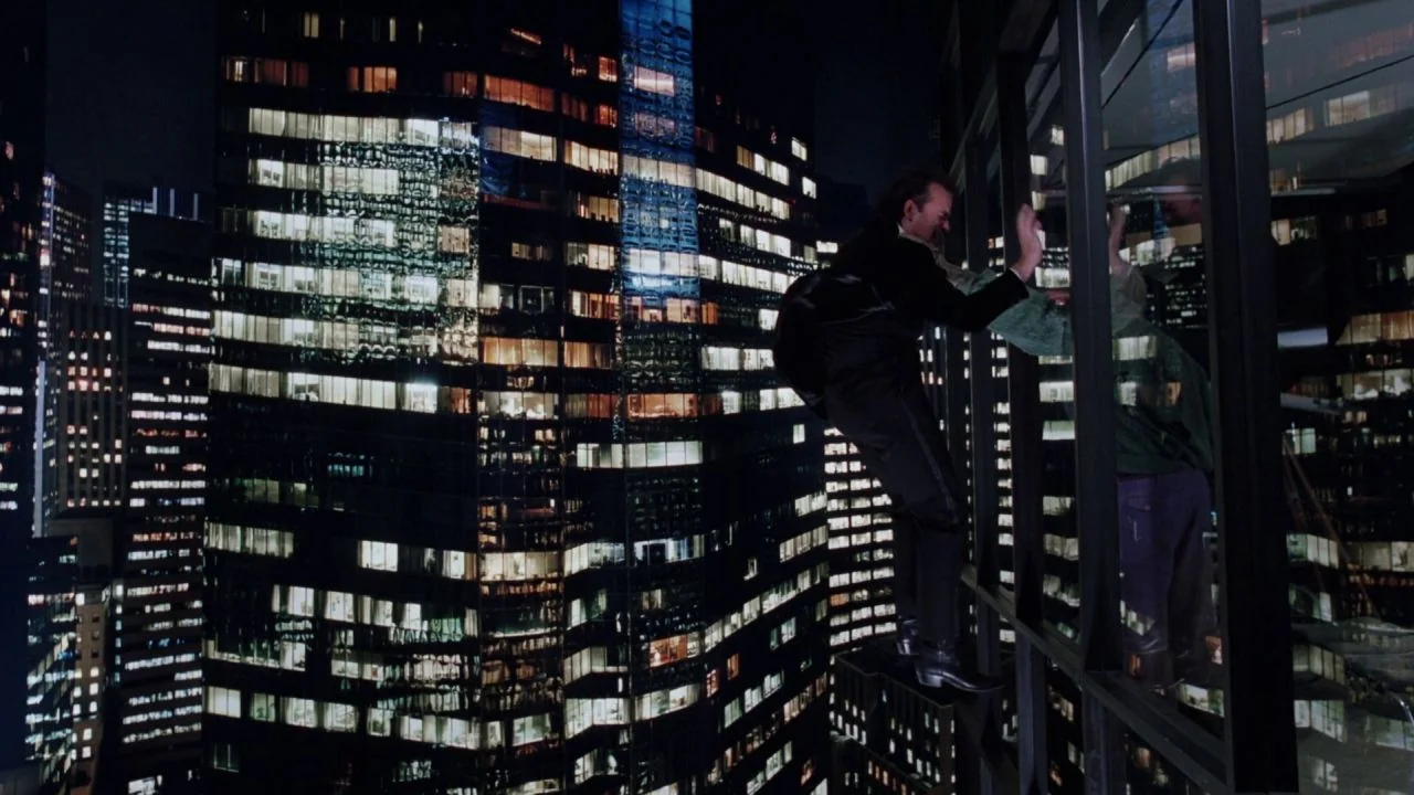 Scrooged (1988) movie still: wide — A high-rise glass facade displays the illuminated grid of a surrounding city skyline at night. A man in a formal…