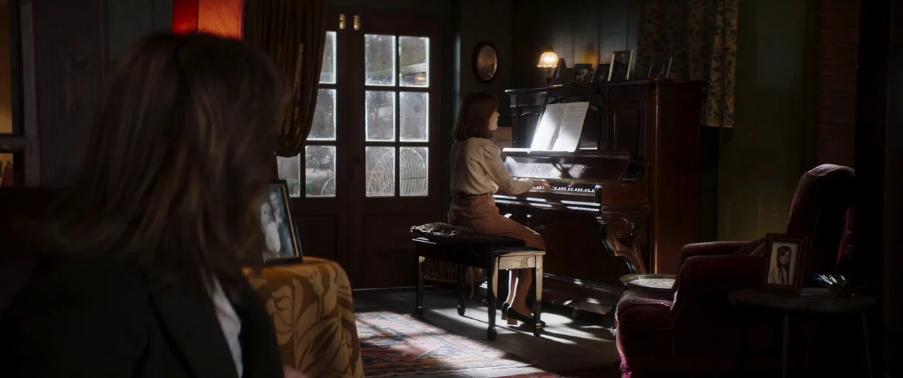 Greta movie still: medium — A furnished interior features an upright piano centered between a large window and a chair. A woman sits on the piano…