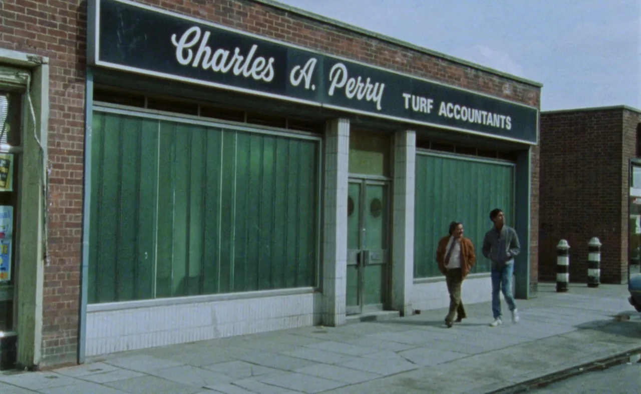 My Beautiful Laundrette (1985) movie still: wide — A brick storefront for a turf accountant features a large sign and glass panels. Two men walk…