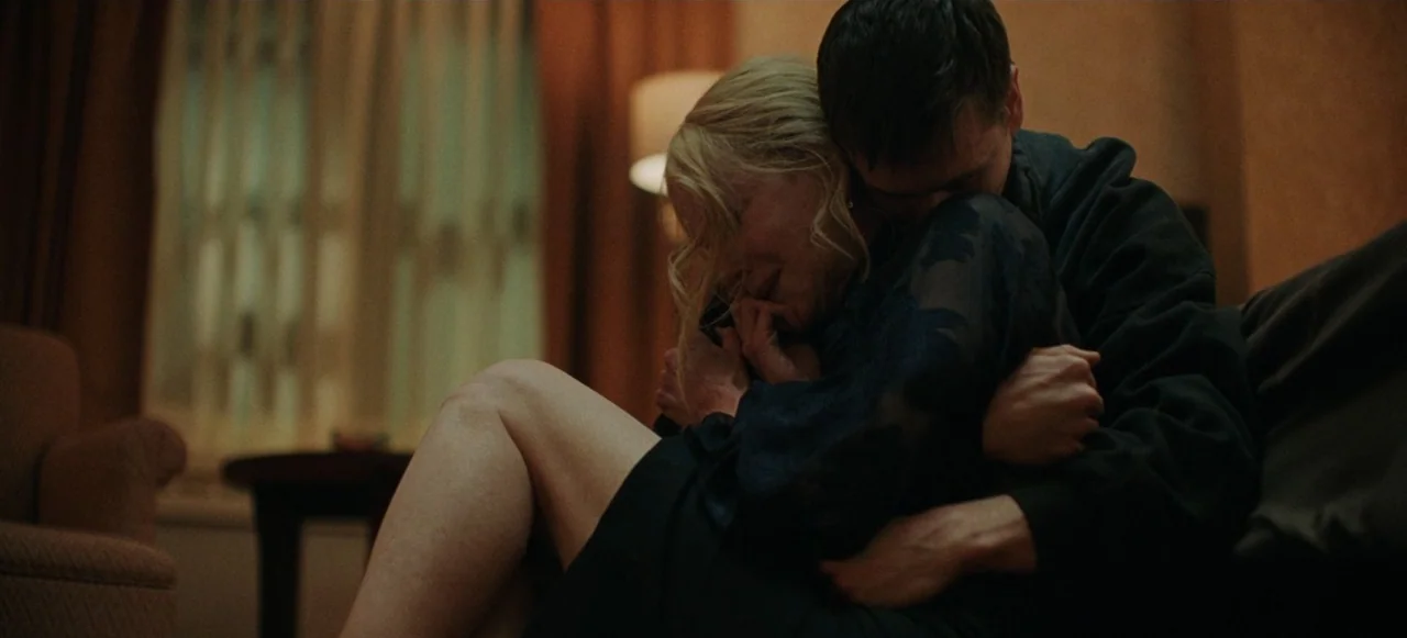 Babygirl (2024) movie still: medium — A dim room contains a beige sofa and a blurred lamp in the background. A man embraces a blonde woman as they…