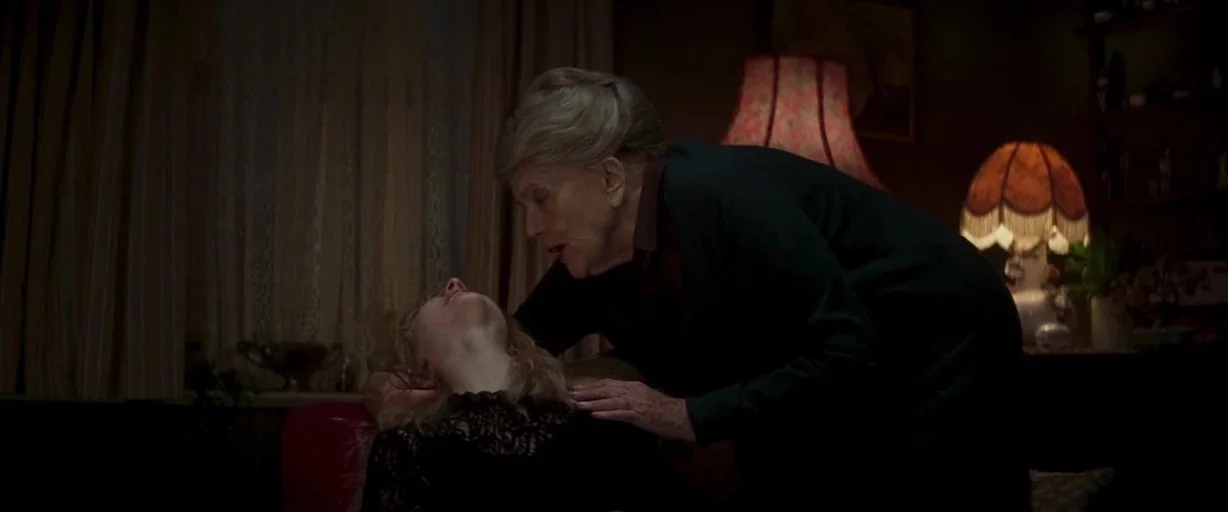 Last Night in Soho (2021) movie still: medium — A dimly lit room features patterned curtains and two ornate vintage lamps. An older woman looks down…