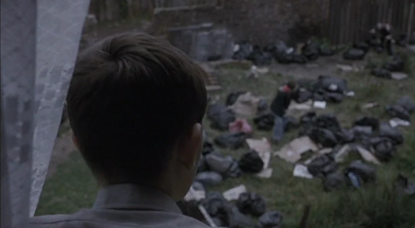 Ratcatcher (1999) movie still: medium — A view from an interior window overlooking a neglected yard covered in refuse. A person stands in the…