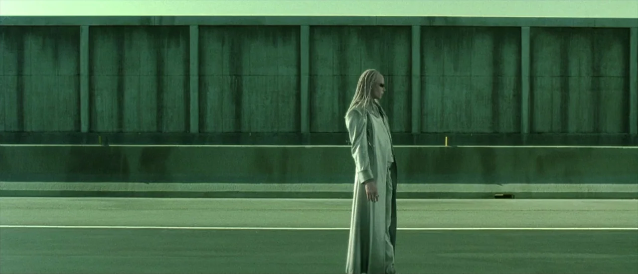 The Matrix Reloaded (2003) movie still: wide — A wide, empty highway stretch features a massive concrete sound barrier wall behind it. A lone figure…