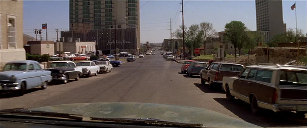 The Getaway (1972) movie still: establishing — An asphalt city street features multiple vintage sedans and station wagons parked along both sides. A…