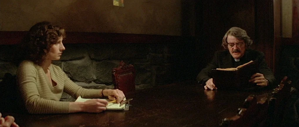 The Fog (1980) movie still: medium — A dimly lit room with stone walls features a long, dark wooden table. A woman sits across from a priest who…