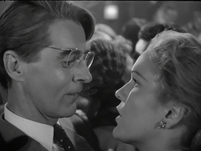A Lesson In Love (1954) movie still: closeup — A crowded, dimly lit indoor social space featuring many blurred figures in the distance. A man and a…
