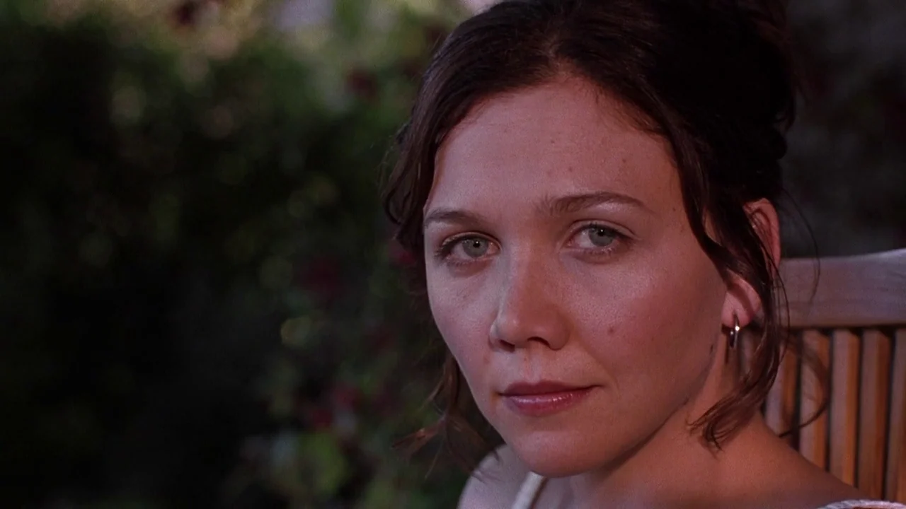 Secretary movie still: closeup — A woman sits outdoors on a wooden chair with foliage blurred in the background. She faces forward and maintains eye…