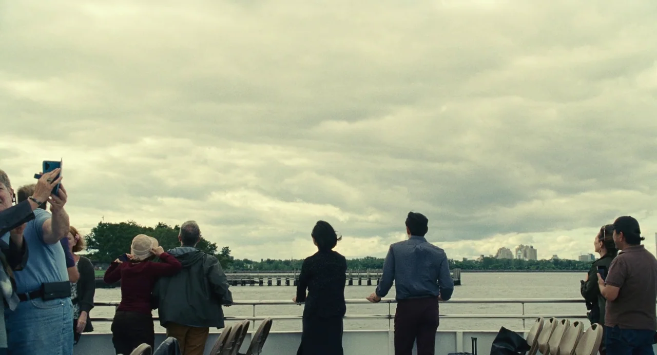 Past Lives (2023) movie still: wide — A group of people stands along a metal railing on an outdoor passenger ferry deck. They look across a wide…