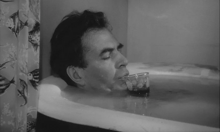 Lolita (1962) movie still: closeup — A man with dark hair rests in a bathtub with a fish-patterned shower curtain visible. He reclines with his eyes…