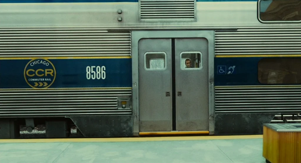 Source Code (2011) movie still: wide — A silver corrugated commuter rail train car sits idle at a concrete station platform. A man looks through the…