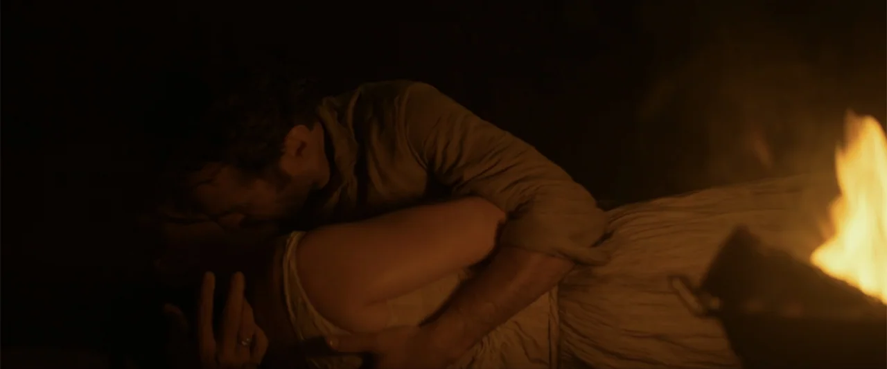 Eiffel (2021) movie still: medium — Two figures lie together in a dark outdoor area near a crackling campfire. The man wraps his arms around the…