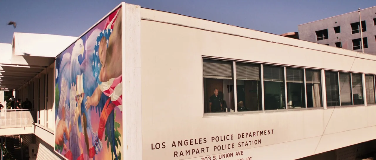 Rampart (2011) movie still: wide — A low-slung, cream-colored concrete building features a large patriotic mural on its corner. The text 'Los…