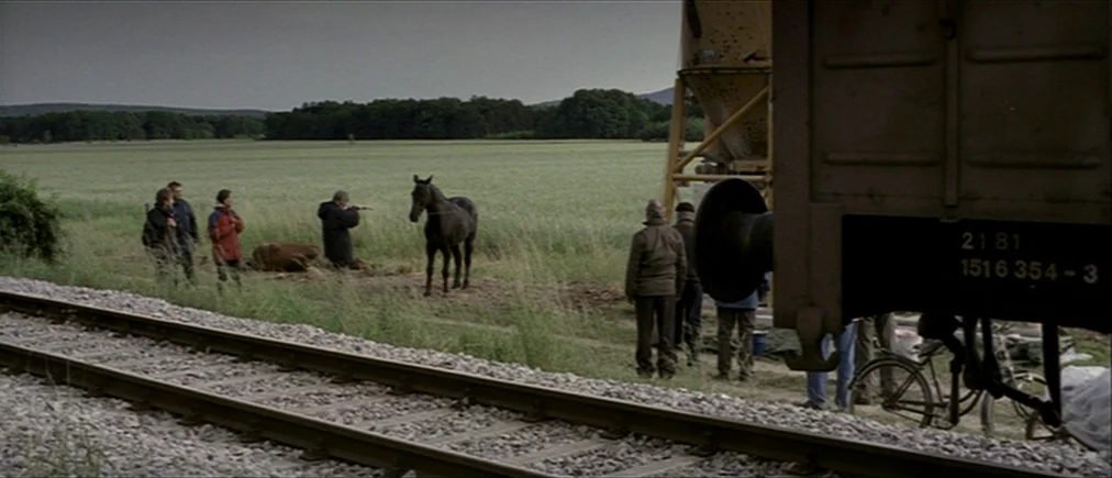 Time Of The Wolf (2003) movie still: wide — A group of people gathers on a grassy strip beside railway tracks in daylight. A dark horse stands near…