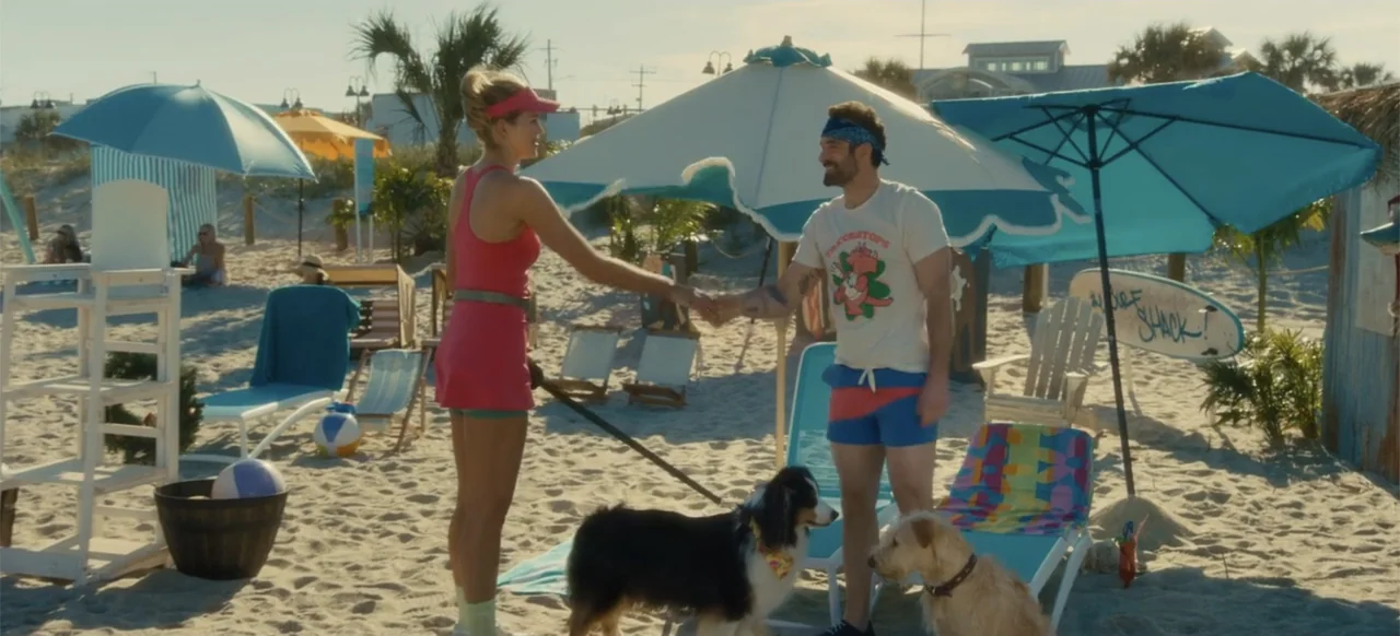 Merv (2025) movie still: fullbody — A sunny sandy beach features colorful umbrellas and lounge chairs under clear blue skies. A woman and a man are…