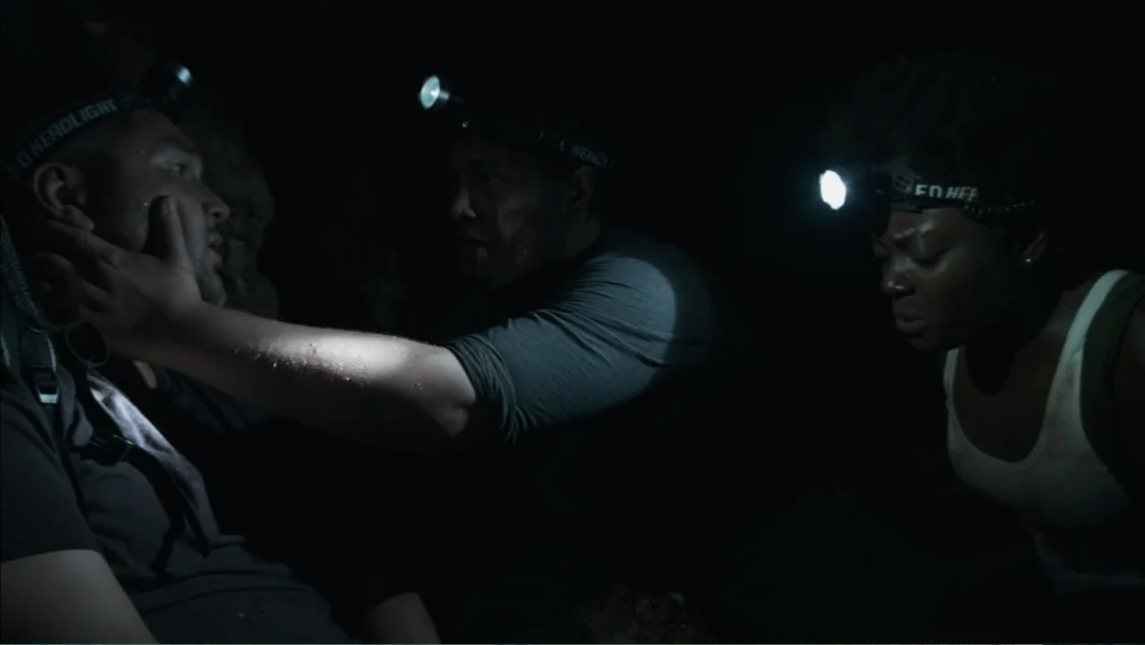 The Grotto (2022) movie still: medium — Three individuals are huddled in a dark, confined space illuminated only by headlamps. One man stabilizes…