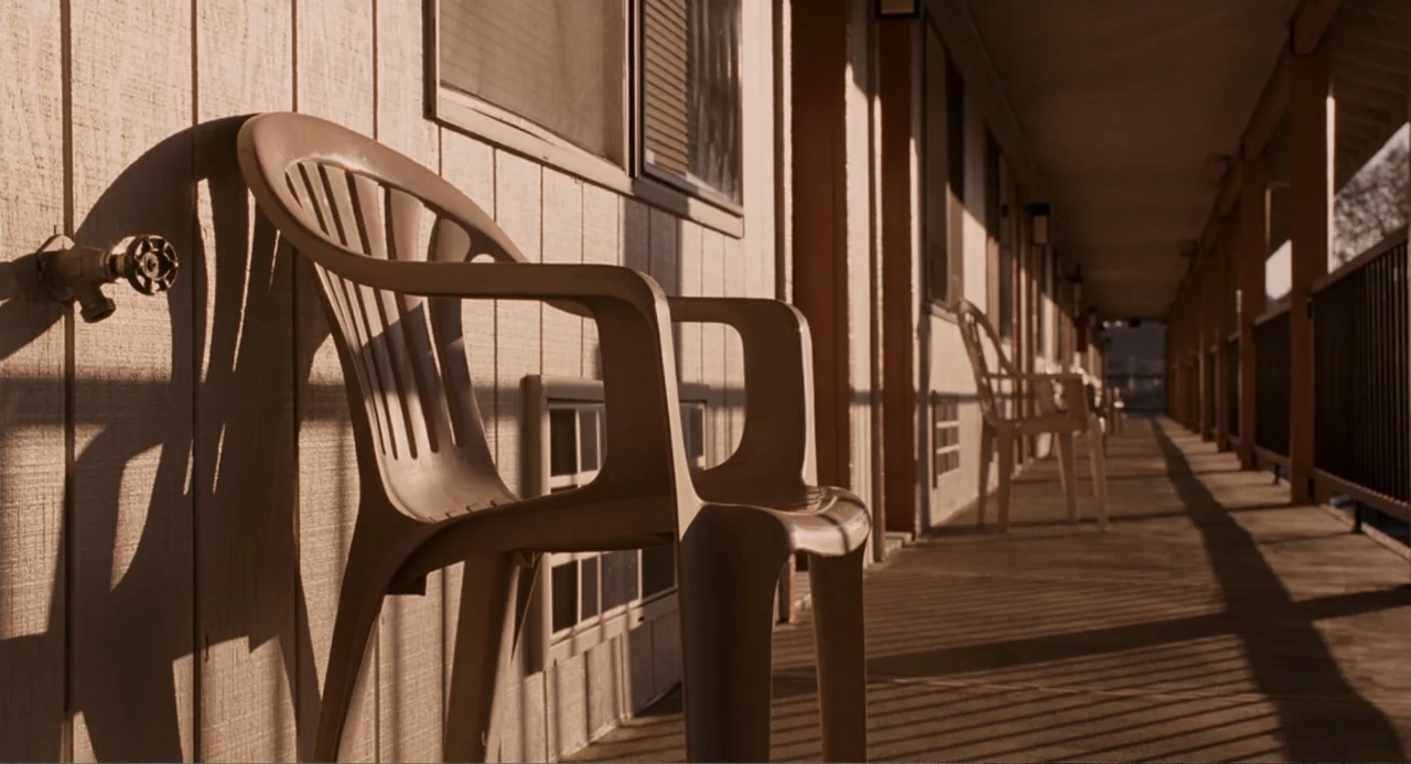 Management (2009) movie still: medium — A wooden motel walkway features empty plastic chairs along a light-colored wall at sunset. Long shadows…