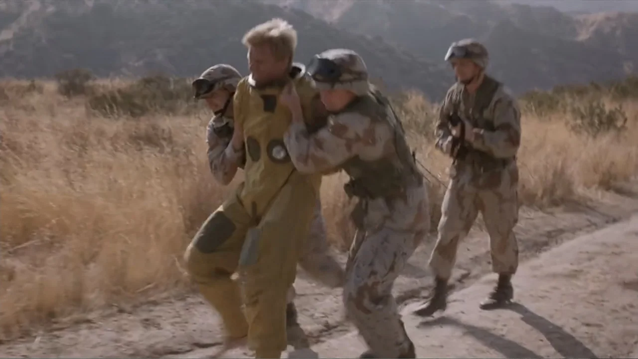 Crisis Point (2012) movie still: fullbody — A dry hilly landscape features a narrow dirt track surrounded by parched yellow grass. Two soldiers in…