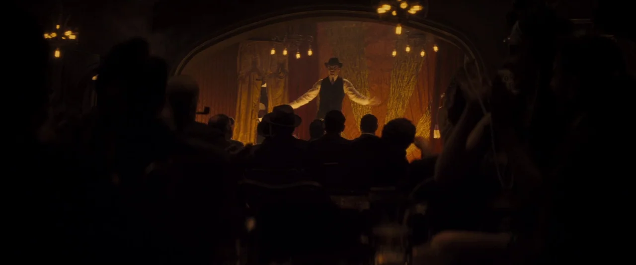 The Immigrant (2013) movie still: wide — A dimly lit vaudeville theater interior features a performer centered on an ornate stage. Silhouetted…