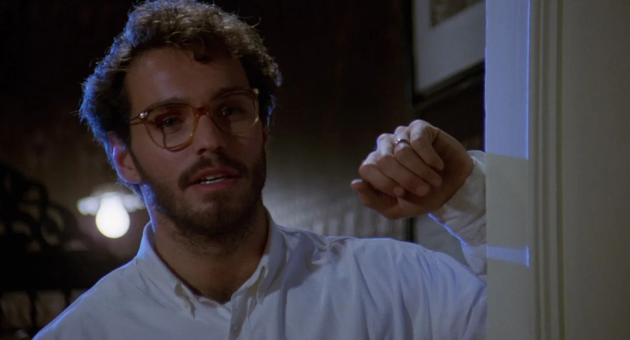 The Spider Labyrinth (1988) movie still: medium — A dark indoor space defined by a wall partition and blurred background. A young man with glasses…