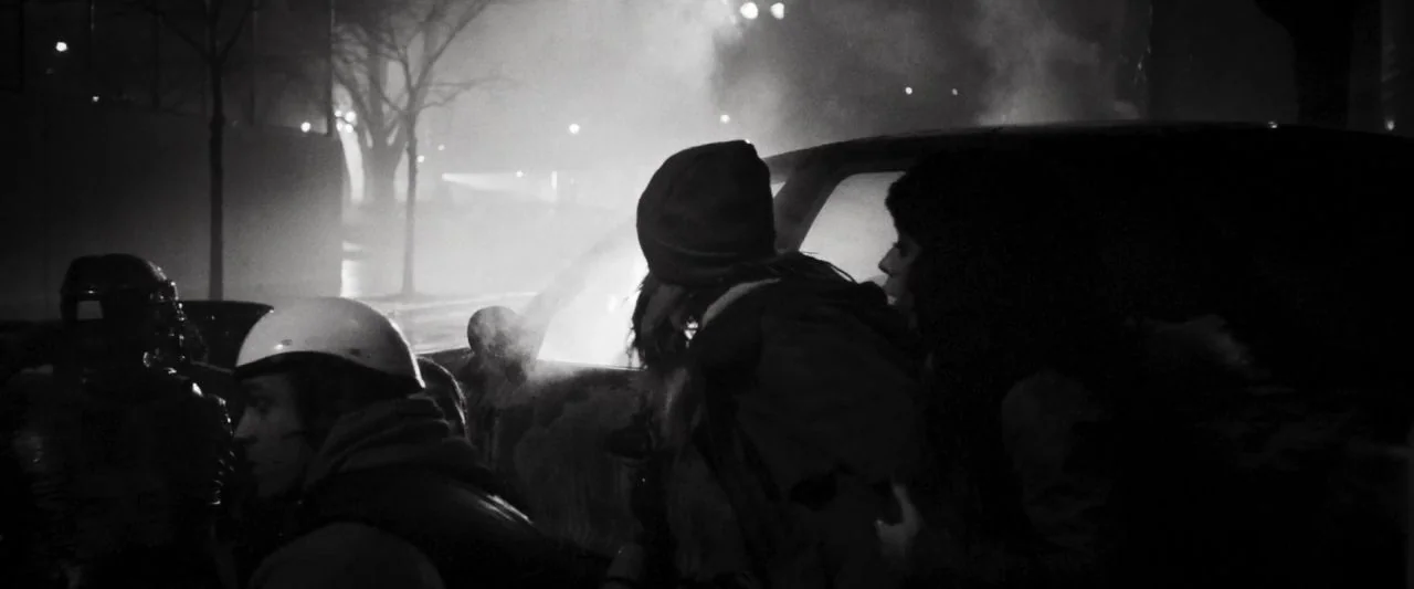 Bushwick (2017) movie still: medium — An urban setting at night features figures standing near a dark vehicle under lights. Thick haze fills the air…