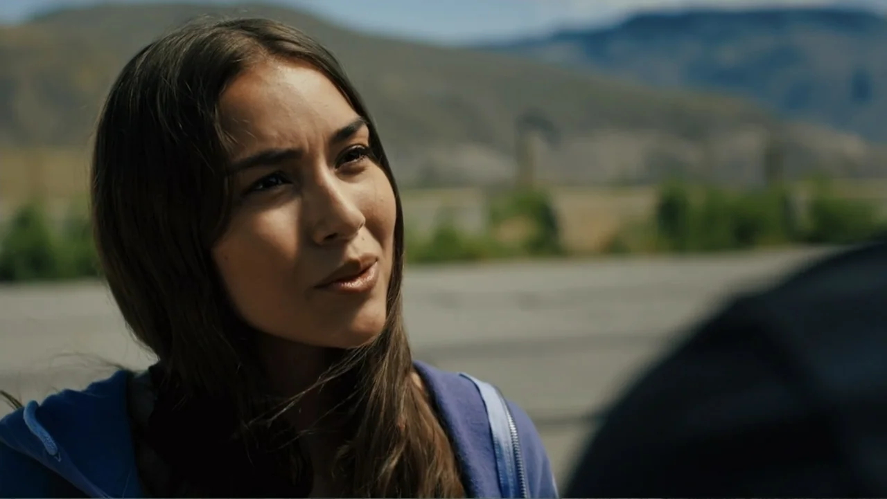 Bullet Proof (2022) movie still: medium — A woman stands in a paved outdoor area with a blurred mountain range background. She listens and speaks to…